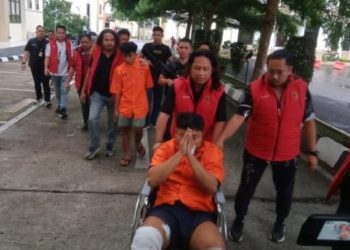 Petugas kepolisian menggiring dua tersangka kasus pembunuhan sopir taksi online Maxim di Jambi. Salah satu tersangka, Hafif (22), menggunakan kursi roda akibat amputasi kaki dan masih menjalani perawatan di RS Bhayangkara Jambi, Selasa (12/8/2025). (Dok. Nahar)